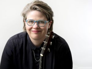 Katie Dahl is smiling facing front at the camera, wearing a black shirt and blue rimmed glasses. She is holding a guitar