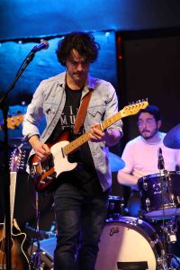 Nicolas is wearing a blue shirt over a black t-shirt with black pants, holding a guitar that is white at the top and black and red at the bottom. He is standing in front of a drummer at his drumset. 