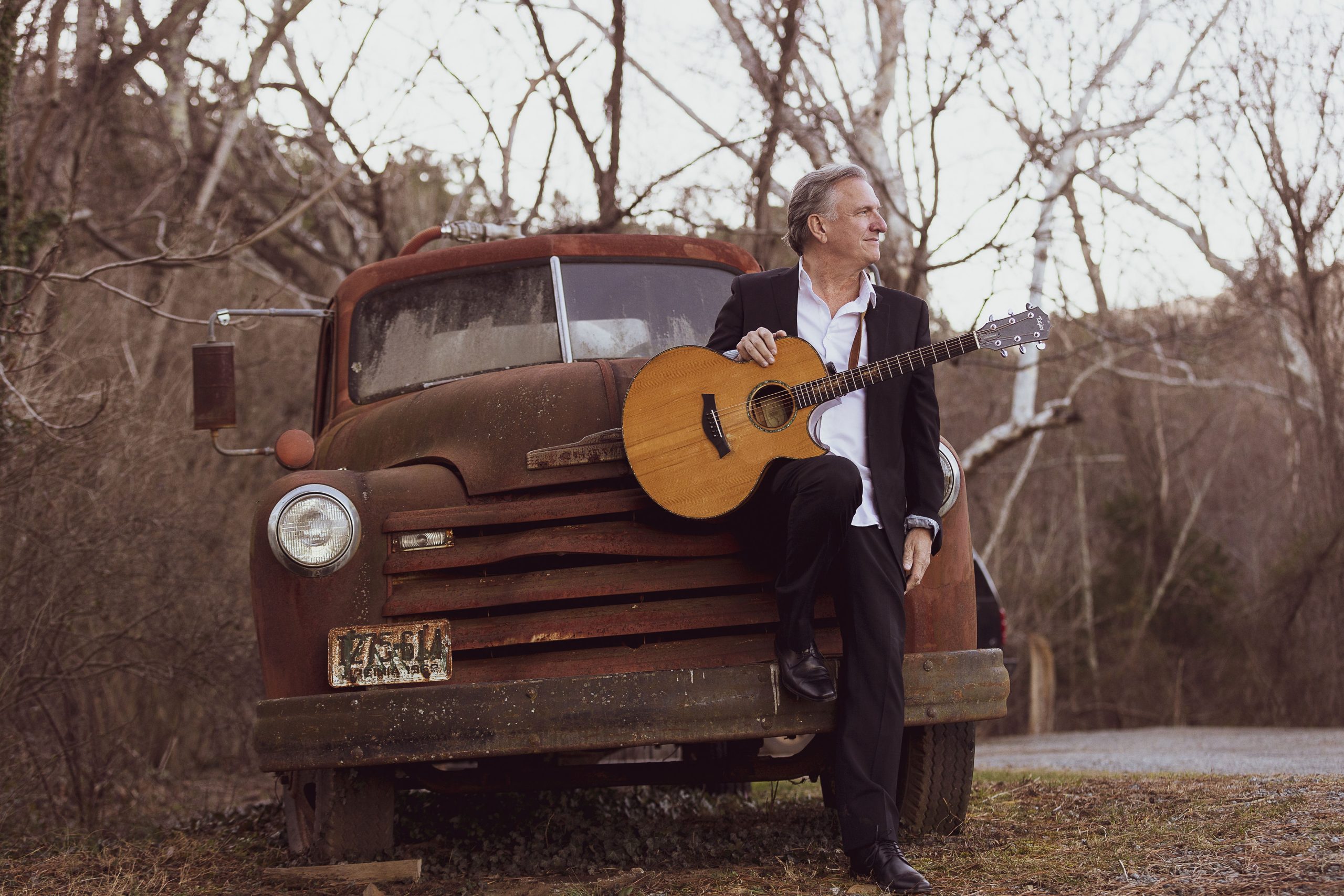 Ellis Paul leaning against an old car with his guitar in an Autumn outdoor setting.