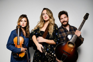photo of trio Barnaby Bright. A woman on the left dressed in a denim shirt, holding a violin, while the middle one has a black dress with yellow flowers and she's holding a guitar. The third on the right wears a brown and black plaid shirt and holds a guitar. 