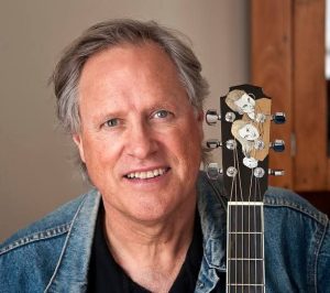 Tom Chapin sits with his guitar by his side, wearing a black t shirt and blue denim jacket.
